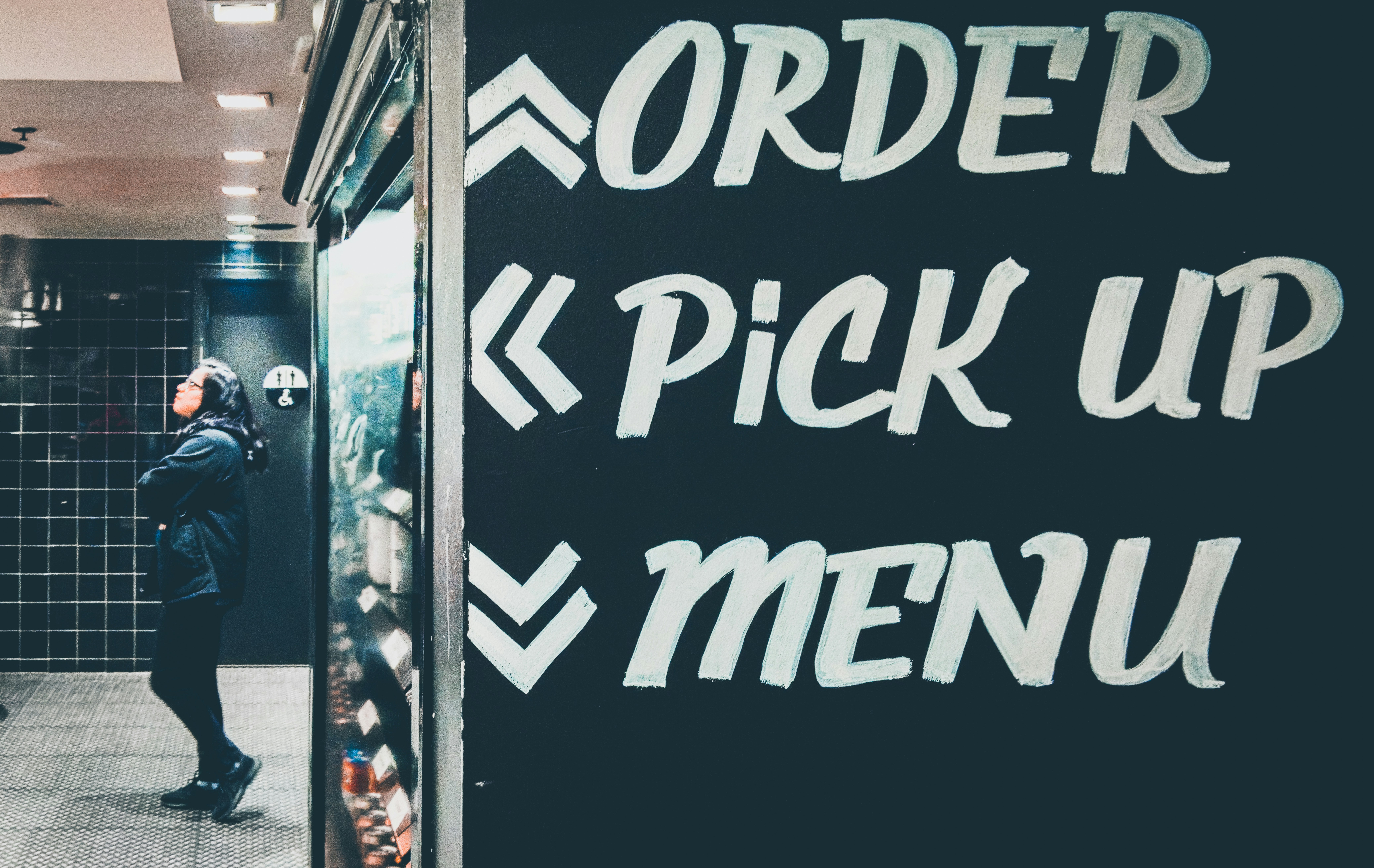 A Hispanic woman wearing a Raiders hoodie and standing in front of user instructions waits to order at And Pizza with a vending machine in the foreground