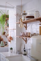 A bright, modern kitchen with white subway tiles on the walls and wooden shelves displaying various kitchen items like bowls, cups, and a kettle. Green plants add a touch of nature, while copper pots and utensils provide a rustic accent. A mix of minimalist and vintage elements creates an inviting and cozy atmosphere.