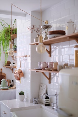 A bright, modern kitchen with white subway tiles on the walls and wooden shelves displaying various kitchen items like bowls, cups, and a kettle. Green plants add a touch of nature, while copper pots and utensils provide a rustic accent. A mix of minimalist and vintage elements creates an inviting and cozy atmosphere.