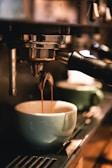 A close-up view of an espresso machine pouring coffee into a light blue cup. The warm lighting creates a cozy atmosphere, highlighting the gleaming metal and the flow of rich coffee.