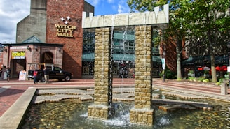 A brick building labeled Witch City Mall is in the background. In the foreground, a fountain with stone pillars and cascading water is surrounded by a small reflective pool. Several people are walking nearby, and a black pickup truck is parked in front. Trees with green foliage are visible, and signage advertising a psychic fair is displayed.