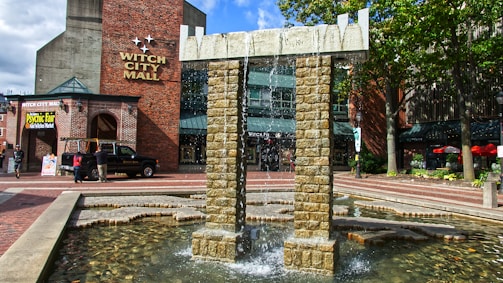 A brick building labeled Witch City Mall is in the background. In the foreground, a fountain with stone pillars and cascading water is surrounded by a small reflective pool. Several people are walking nearby, and a black pickup truck is parked in front. Trees with green foliage are visible, and signage advertising a psychic fair is displayed.