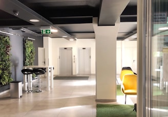A modern interior space featuring two elevators at the back, surrounded by white walls and a ceiling with black beams. On the left, there is a tall table with two black bar stools and a trash can. Vertical green plant walls add a touch of nature. To the right, there are yellow chairs on a patch of green carpet.