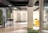 A modern interior space featuring two elevators at the back, surrounded by white walls and a ceiling with black beams. On the left, there is a tall table with two black bar stools and a trash can. Vertical green plant walls add a touch of nature. To the right, there are yellow chairs on a patch of green carpet.