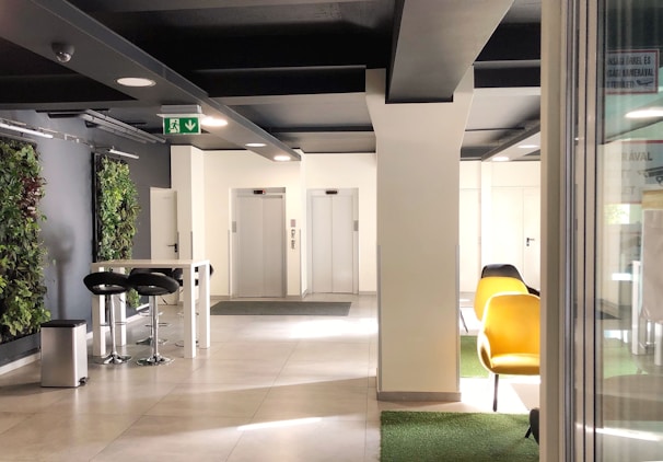 A modern interior space featuring two elevators at the back, surrounded by white walls and a ceiling with black beams. On the left, there is a tall table with two black bar stools and a trash can. Vertical green plant walls add a touch of nature. To the right, there are yellow chairs on a patch of green carpet.