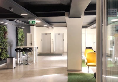 A modern interior space featuring two elevators at the back, surrounded by white walls and a ceiling with black beams. On the left, there is a tall table with two black bar stools and a trash can. Vertical green plant walls add a touch of nature. To the right, there are yellow chairs on a patch of green carpet.