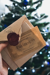 A hand is holding a ticket and an envelope with a wax seal in front of a blurred Christmas tree. The ticket reads 'Hogwarts Express' and 'Platform 9 3/4', evoking a magical theme.