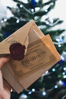 A hand is holding a ticket and an envelope with a wax seal in front of a blurred Christmas tree. The ticket reads 'Hogwarts Express' and 'Platform 9 3/4', evoking a magical theme.