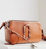 A stylish brown leather handbag with a glossy metallic clasp prominently displayed. The design is sleek and compact, with a long strap and detailed stitching. The bag is positioned on a minimalistic white surface, enhancing its elegant appearance.
