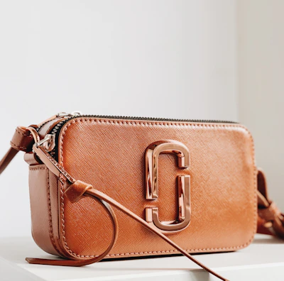 A stylish brown leather handbag with a glossy metallic clasp prominently displayed. The design is sleek and compact, with a long strap and detailed stitching. The bag is positioned on a minimalistic white surface, enhancing its elegant appearance.