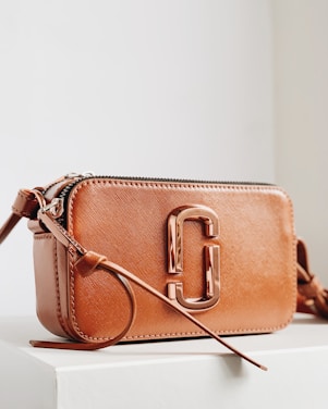 A stylish brown leather handbag with a glossy metallic clasp prominently displayed. The design is sleek and compact, with a long strap and detailed stitching. The bag is positioned on a minimalistic white surface, enhancing its elegant appearance.