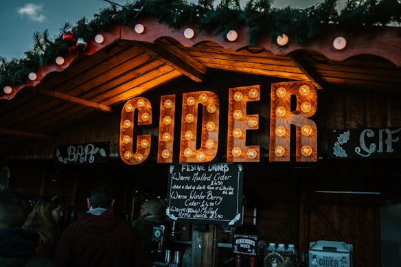 An outdoor market stall features a large, illuminated sign spelling 'CIDER' with warm, decorative lights. The stall is decorated with festive greenery, including holly and Christmas ornaments. Below the sign, a menu board lists various festive drink options like warm mulled cider. A small crowd gathers, and bottles and jugs can be seen on display.