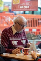 An elderly man wearing glasses and a maroon sweater with polar bear patterns sits at a wooden table, reading a newspaper. In front of him are paper cups with a recognizable coffee brand logo, and there are some plastic utensils and lids on the table. The scene is indoors, with a background of an orange construction barrier and blurred outdoor elements visible through the window.