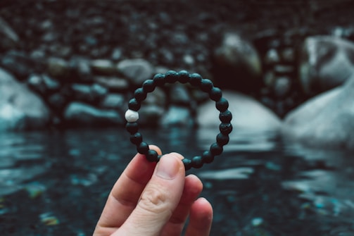 A hand holding a bracelet made of multiple dark beads and one white bead is in focus. The background shows a natural scene with water and rocks, creating a tranquil and serene atmosphere.