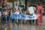 A group of people is marching on a city street, holding a large banner that reads 'Tell the Truth.' The participants appear to be part of a protest or demonstration and are dressed in casual clothing, with some wearing lab coats. A red telephone booth and a tree are visible in the background, and the scene is wet, likely due to rain.