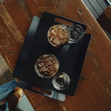 A top-down view of a black tray on a wooden table with two glasses of iced coffee and two glasses of water. The glasses contain ice cubes and straws are present in the iced coffee.