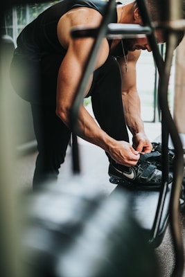 A muscular person is adjusting their athletic shoes while seated on a bench or similar surface in a gym environment. The individual is wearing a sleeveless top and athletic leggings. The gym appears to have natural lighting streaming in from windows, creating a focused and intense atmosphere.