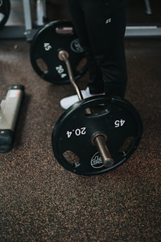 A barbell with 45-pound weight plates rests on a speckled gym floor. A person wearing black pants and white sneakers stands nearby, partially visible.