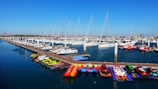 Yachts and sailboats are docked in a marina with a wooden pier extending into the water. Various jet skis and inflatable boats are lined up along the dock. The sky is clear and blue, creating a serene backdrop for the scene.