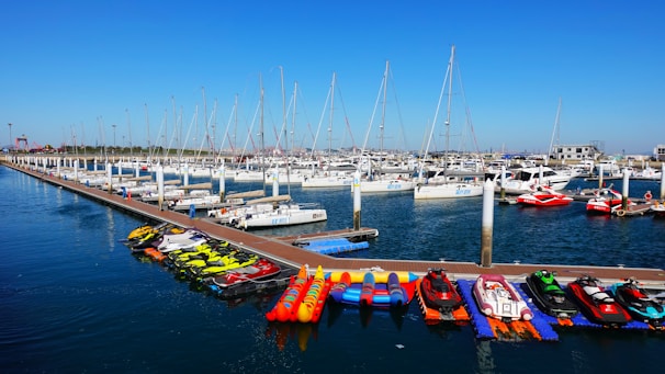 Yachts and sailboats are docked in a marina with a wooden pier extending into the water. Various jet skis and inflatable boats are lined up along the dock. The sky is clear and blue, creating a serene backdrop for the scene.