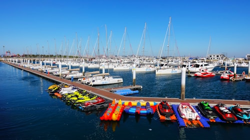 Yachts and sailboats are docked in a marina with a wooden pier extending into the water. Various jet skis and inflatable boats are lined up along the dock. The sky is clear and blue, creating a serene backdrop for the scene.