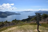 A scenic viewpoint overlooks a vast body of water with several islands and mountainous terrain covered in lush greenery. In the foreground, there is a multi-directional wooden signpost with arrows pointing to various city names like New York, Auckland, and Paris. The sky is mostly clear with some scattered clouds.