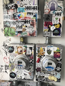 The image features two utility meters, each covered with a diverse array of colorful stickers and graffiti art on a rough, painted wall. The stickers include various designs, such as logos, text, drawings, and symbols, showcasing an urban, street-art aesthetic.