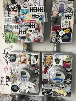 The image features two utility meters, each covered with a diverse array of colorful stickers and graffiti art on a rough, painted wall. The stickers include various designs, such as logos, text, drawings, and symbols, showcasing an urban, street-art aesthetic.