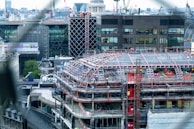 The image depicts a construction site of a large building in an urban area. A partially completed structure with scaffolding covers the upper sections, and workers in safety gear are visible on various levels. Surrounding the site are modern office buildings, some with reflective glass facades. Cranes and other construction equipment are also present in the vicinity.