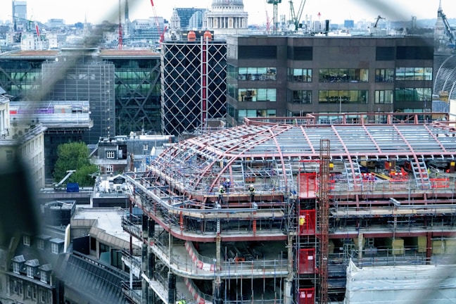 The image depicts a construction site of a large building in an urban area. A partially completed structure with scaffolding covers the upper sections, and workers in safety gear are visible on various levels. Surrounding the site are modern office buildings, some with reflective glass facades. Cranes and other construction equipment are also present in the vicinity.