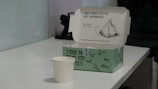 An open takeaway box on a table, with the lid displaying a sketch of a samosa and the text 'Now you'll never get confused! Your samosa has a name. Only the freshest samosa delivered to you!' The box is green with additional text and graphics. In front of the box, there is a white paper cup. The setting appears to be an office or indoor environment with a white surface and a telephone blurred in the background.