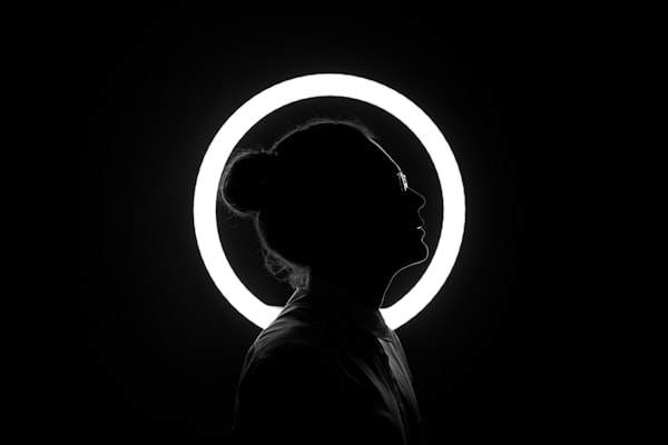 a figure with hair in a bun and glasses, their head surrounded by a circle of light, against a black background