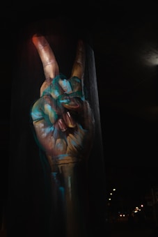A large, detailed mural of a human hand making a peace sign is painted on a cylindrical urban structure. The hand is artistically highlighted with blue and orange shades, standing out against the dark background. Streetlights and distant city lights create a nighttime urban atmosphere.