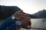 A close-up of a traveler’s hand adjusting a high-quality watch against a backdrop of mountain peaks.