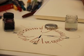 A detailed lettering piece is crafted on a white sheet, resembling a clock face with elaborate calligraphy. There are ink jars and tools scattered nearby on a desk, including one with black ink and another containing clear liquid. Pens and markers are also visible in the background, adding to the artistic setting.