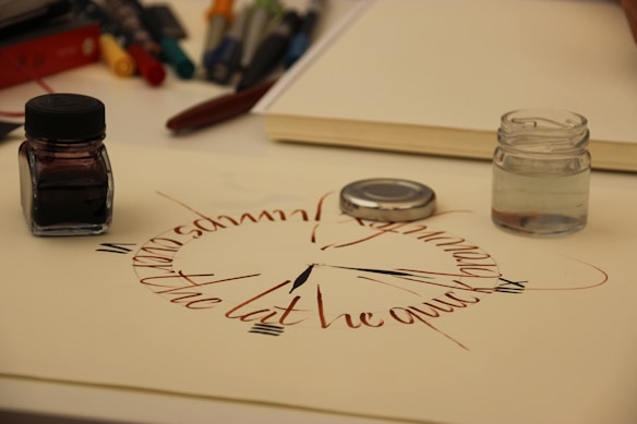 A detailed lettering piece is crafted on a white sheet, resembling a clock face with elaborate calligraphy. There are ink jars and tools scattered nearby on a desk, including one with black ink and another containing clear liquid. Pens and markers are also visible in the background, adding to the artistic setting.