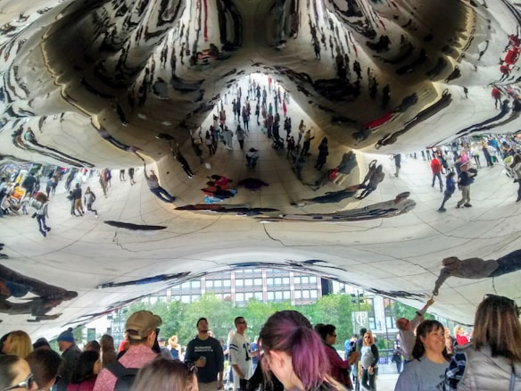 A large reflective sculpture creates distorted reflections of numerous people gathered underneath it. The reflections include figures of people who are strolling, standing, or interacting with the sculpture. The surface captures the reflections in a chaotic, abstract manner, emphasizing the structure's unique shape and material.