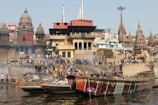 People gather along a bustling riverside ghat surrounded by historic and religious architecture. Smoke rises from burning pyres, suggesting cremation activity. Several boats are floating on the river, and the area is filled with a sense of cultural and spiritual significance.