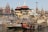 People gather along a bustling riverside ghat surrounded by historic and religious architecture. Smoke rises from burning pyres, suggesting cremation activity. Several boats are floating on the river, and the area is filled with a sense of cultural and spiritual significance.