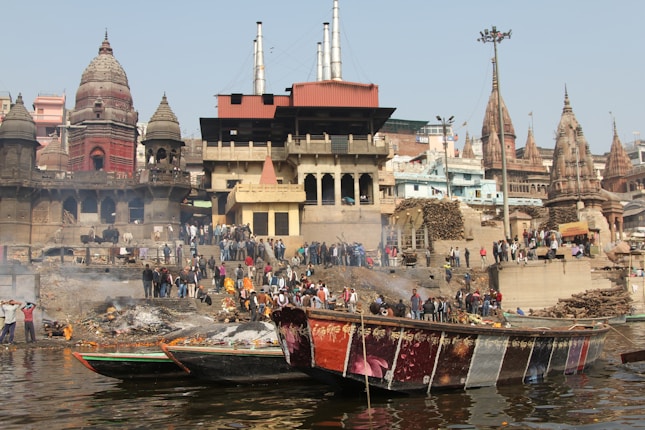 People gather along a bustling riverside ghat surrounded by historic and religious architecture. Smoke rises from burning pyres, suggesting cremation activity. Several boats are floating on the river, and the area is filled with a sense of cultural and spiritual significance.