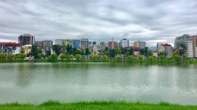 A lakeside cityscape with a view of modern and traditionally styled buildings. The scene includes a body of water in the foreground, lined with lush green grass. Trees and vegetation are visible between the lake and the buildings, which vary in color and height. The sky is overcast, casting soft reflections on the water.
