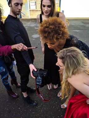 A group of people is gathered around, looking intently at the screen of a professional camera held by one individual. They appear to be reviewing photographs, possibly from a photo shoot. The setting is outdoors, with a concrete ground and a building wall in the background.