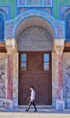 A person walks in front of a richly decorated building entrance with intricate tilework featuring geometric patterns and Arabic calligraphy. The structure has ornate columns with a mix of blue, green, and red hues.