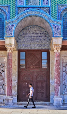 A person walks in front of a richly decorated building entrance with intricate tilework featuring geometric patterns and Arabic calligraphy. The structure has ornate columns with a mix of blue, green, and red hues.