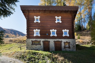 A charming, rustic wooden house with distinct white window frames and ornate architectural features, situated in a serene natural setting surrounded by trees and distant hills.