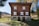 A charming, rustic wooden house with distinct white window frames and ornate architectural features, situated in a serene natural setting surrounded by trees and distant hills.