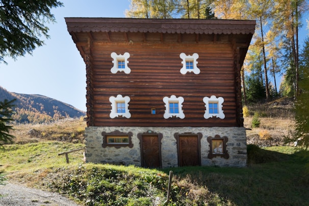 A charming, rustic wooden house with distinct white window frames and ornate architectural features, situated in a serene natural setting surrounded by trees and distant hills.