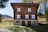 A charming, rustic wooden house with distinct white window frames and ornate architectural features, situated in a serene natural setting surrounded by trees and distant hills.