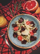 A plate of salad featuring slices of cured meat, cherry tomatoes, and shavings of parmesan cheese, set on a blue ceramic dish. The table is adorned with a red and black checkered cloth, sprigs of rosemary, a halved grapefruit, and a glass of orange juice are also present.
