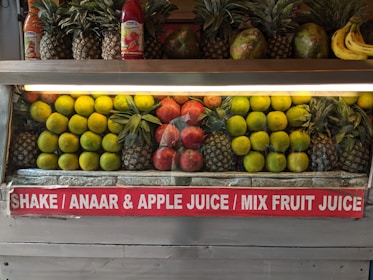 A fruit display featuring neatly arranged rows of pineapples, citrus fruit, pomegranates, and a few bananas on the side. Two juice bottles are also visible on the shelf. A sign below advertises various fruit juices like Shake, Anaar (pomegranate), Apple Juice, and Mix Fruit Juice.